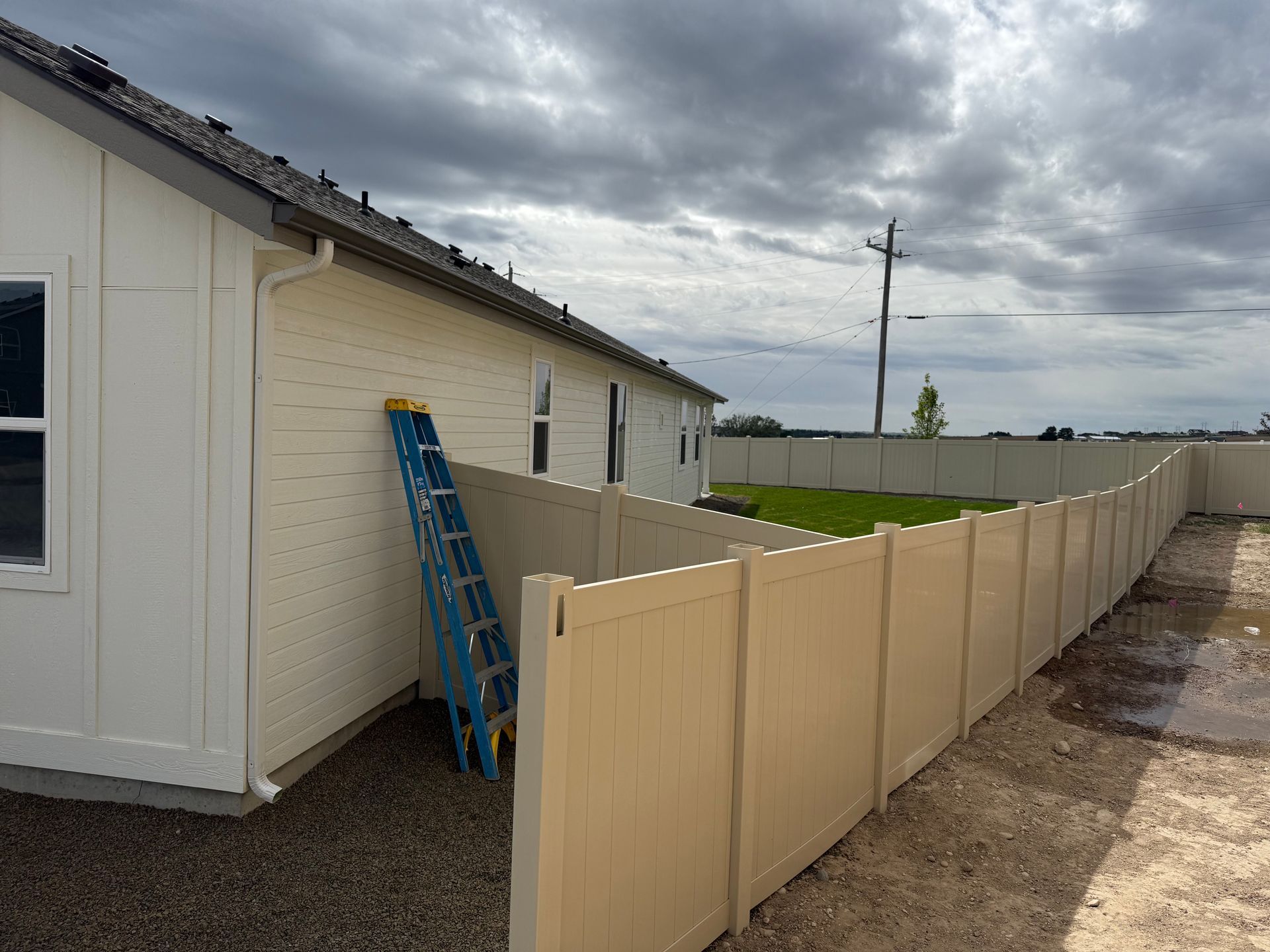 Beige vinyl fence surrounds a house with a blue ladder leaning against the cream-colored siding under a cloudy sky.
