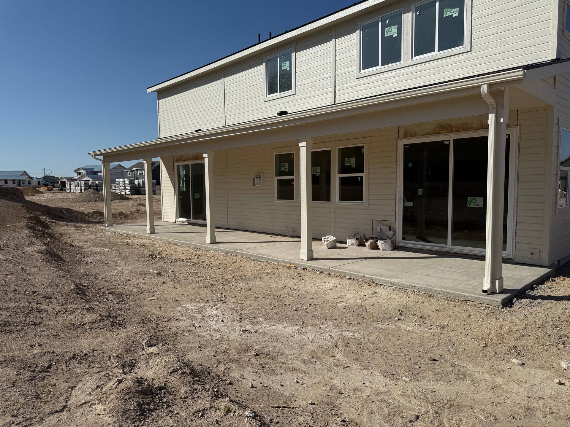 Back of a two-story beige house under construction with a long concrete patio. Dirt foreground, blue sky.