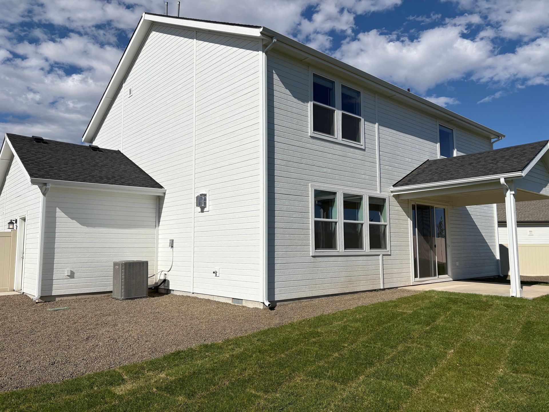 White two-story house with attached garage and patio. Green grass in the foreground, gravel on the side. Blue sky.