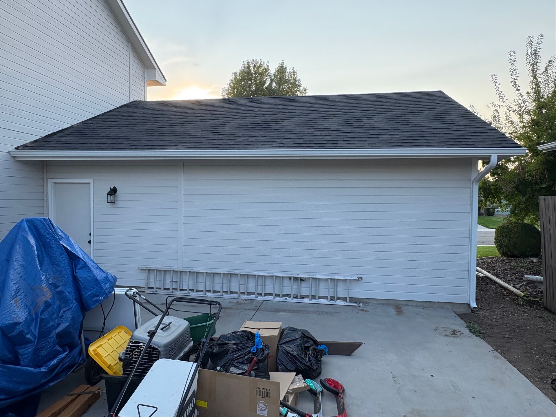 A white-sided garage with a black roof. Clutter is in front, a blue tarp is on the left.