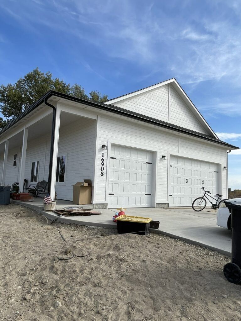 White house with black trim, two garage doors, porch, blue sky. Gravel and concrete yard with a bike.