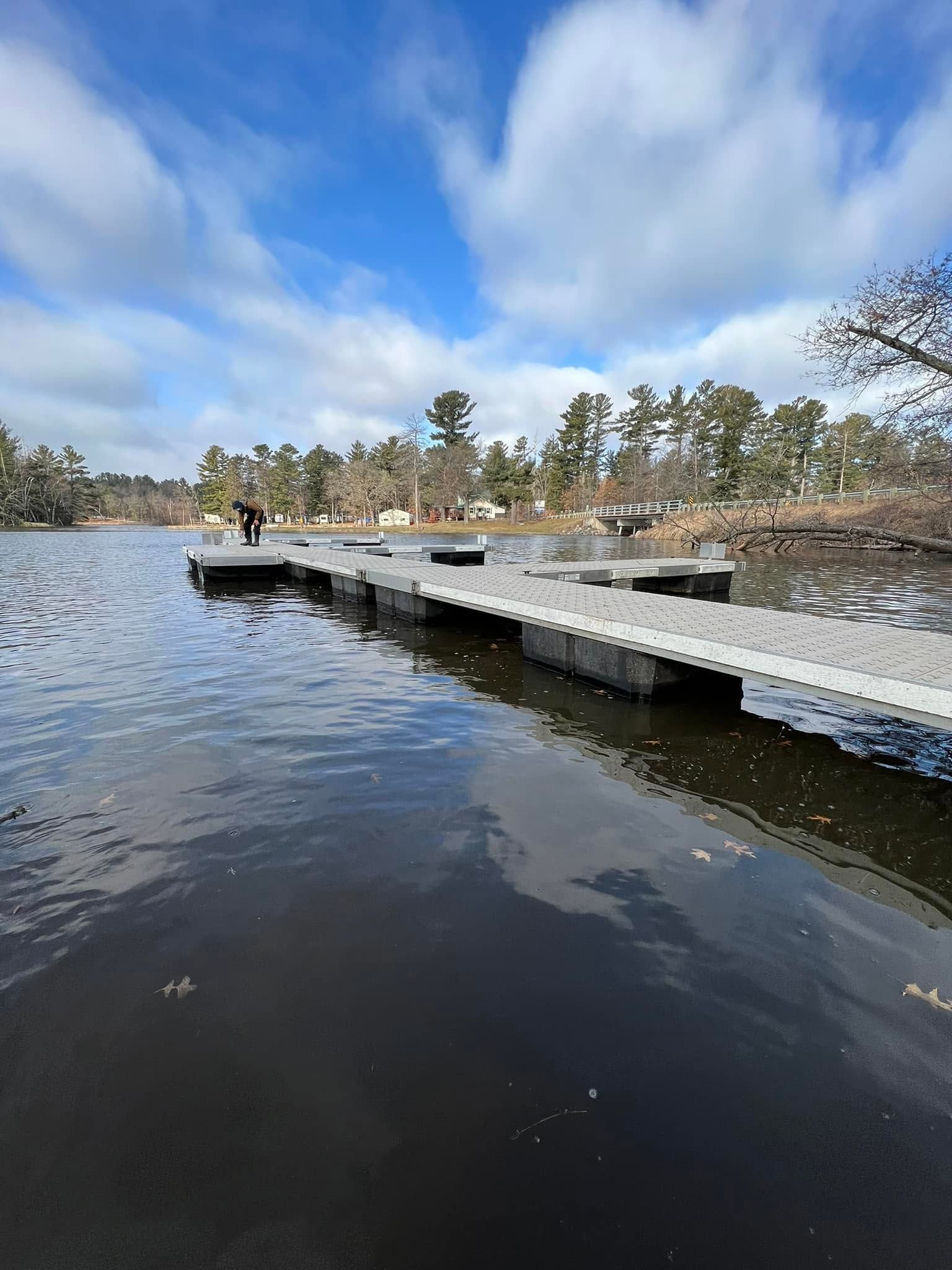 Wooden dock extending over a calm lake under a partly cloudy blue sky, with trees lining the shore.
