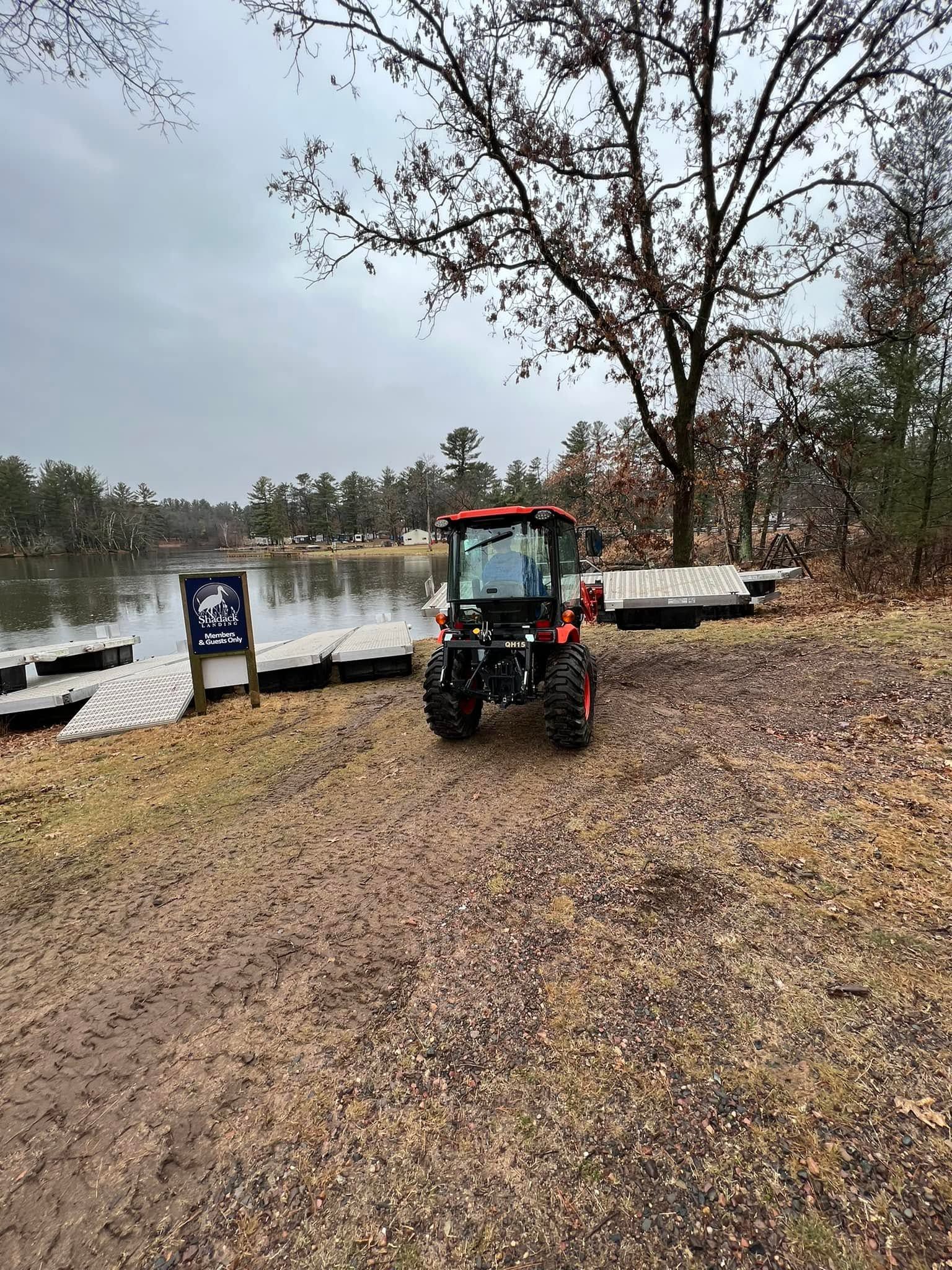 Red tractor on a gravel path beside a lake under an overcast sky, with a dock and bare trees nearby