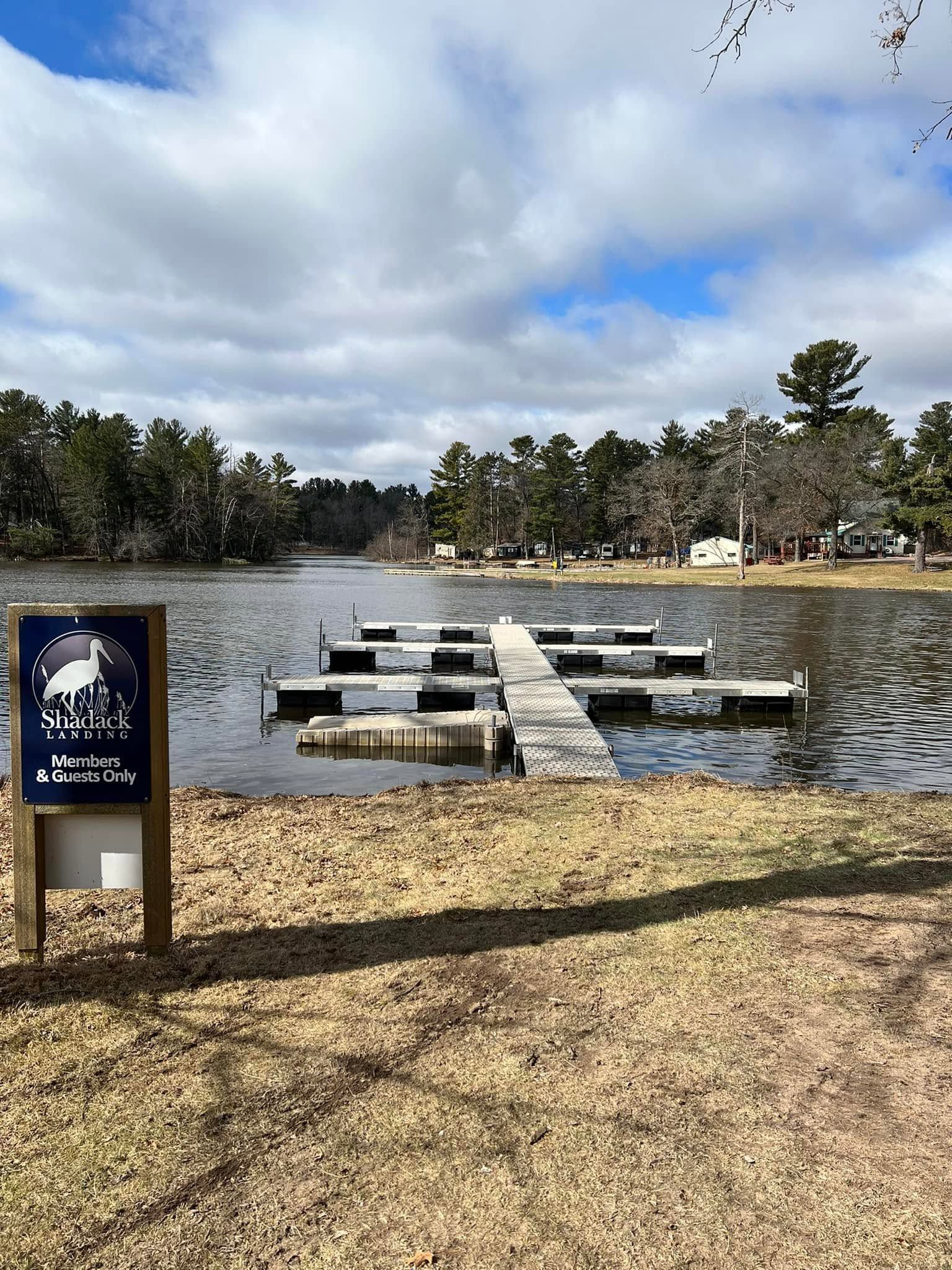 Floating docks on a small lake beside a sandy shore, with a sign in the foreground and cloudy sky.