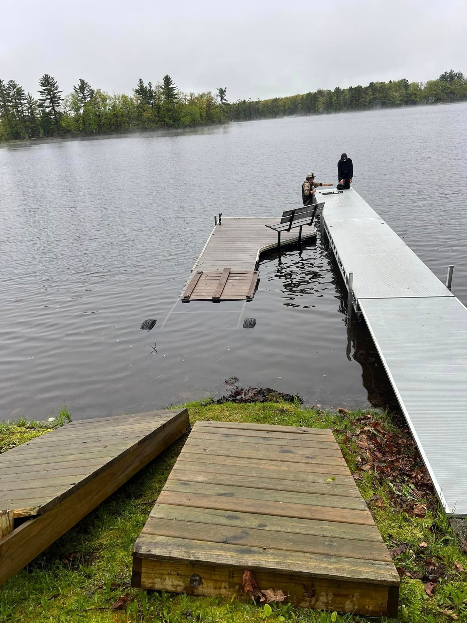 Wooden dock extending into a calm lake, with two people standing near the end and a small platform nearby