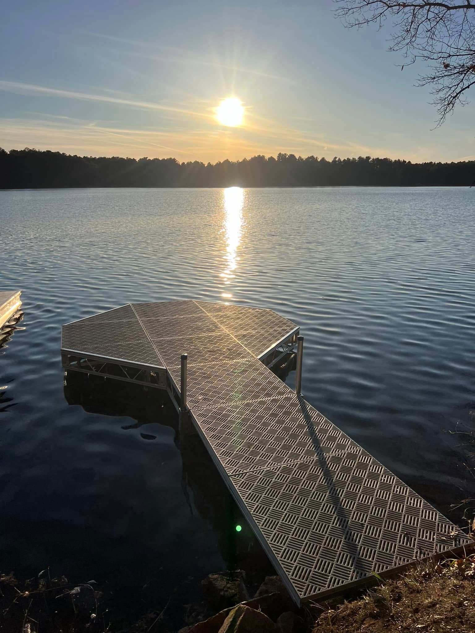 Sunset over a calm lake, with a small dock in the foreground and sunlight reflecting on the water