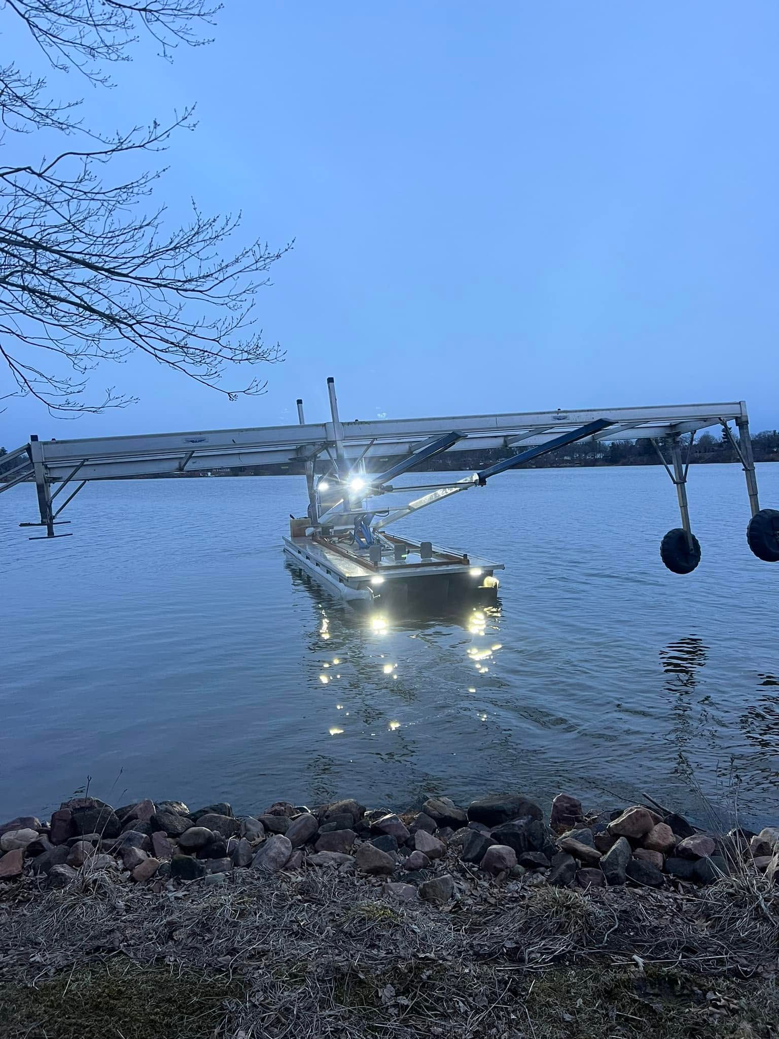 Small boat with bright headlights on calm blue water at dusk, near a rocky shoreline and bare branches.