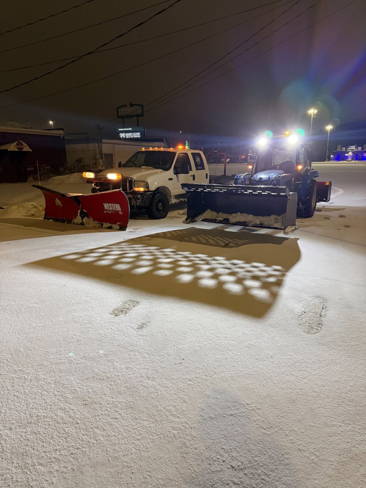 Snowplow with bright headlights and plow blade clearing a snowy lot at night