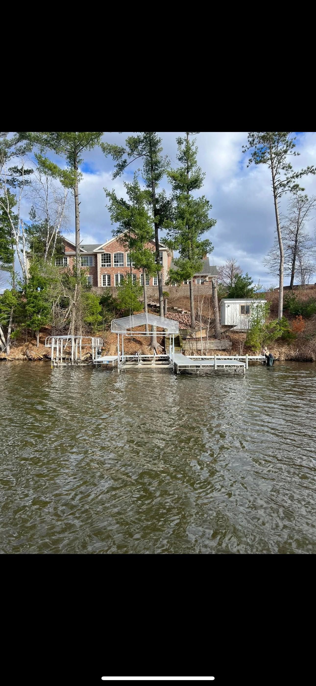 A lake with rippling water, docks, and a house among tall trees under a partly cloudy sky.