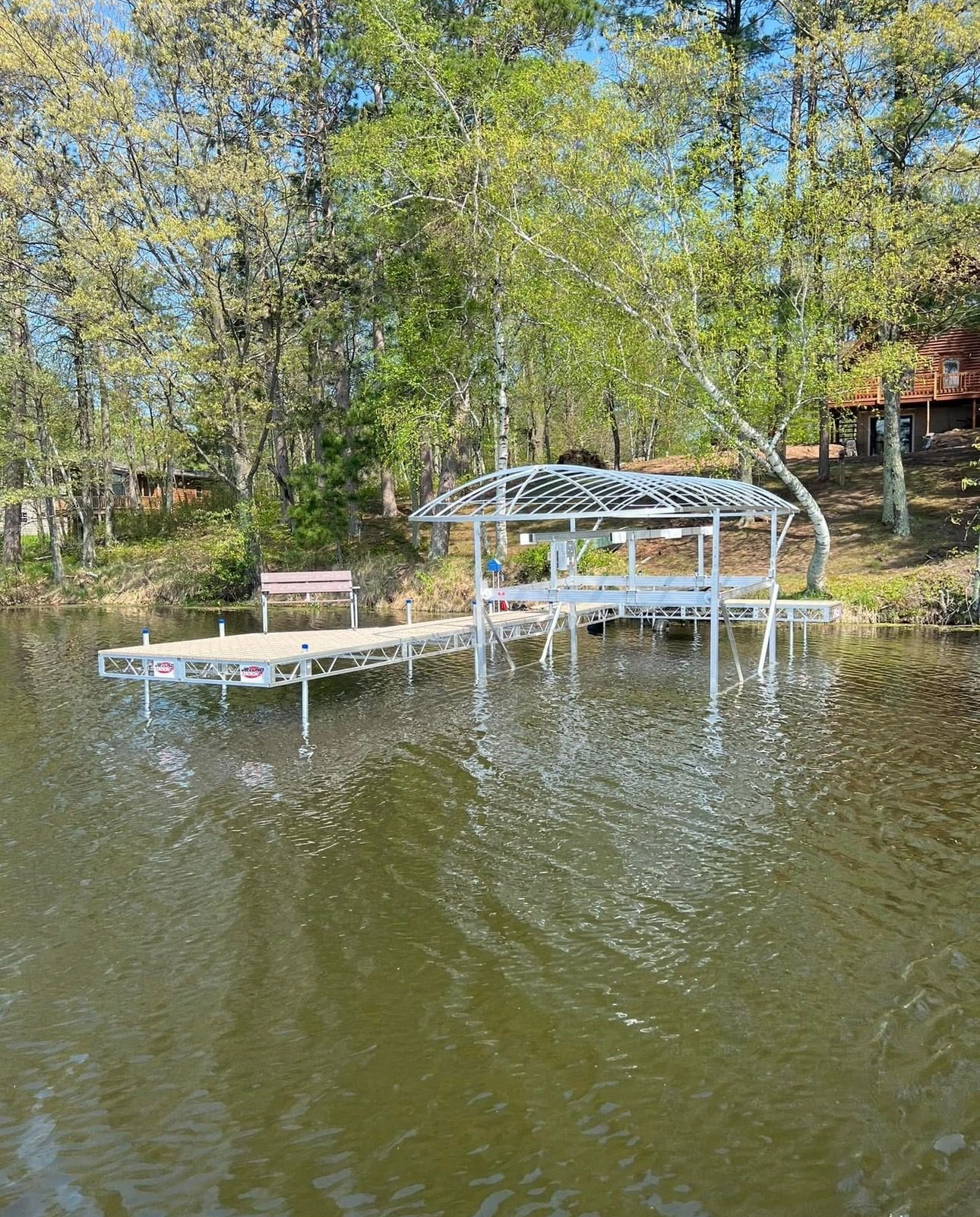 Wooden dock with a bench and canopy on a calm lake, surrounded by spring trees.