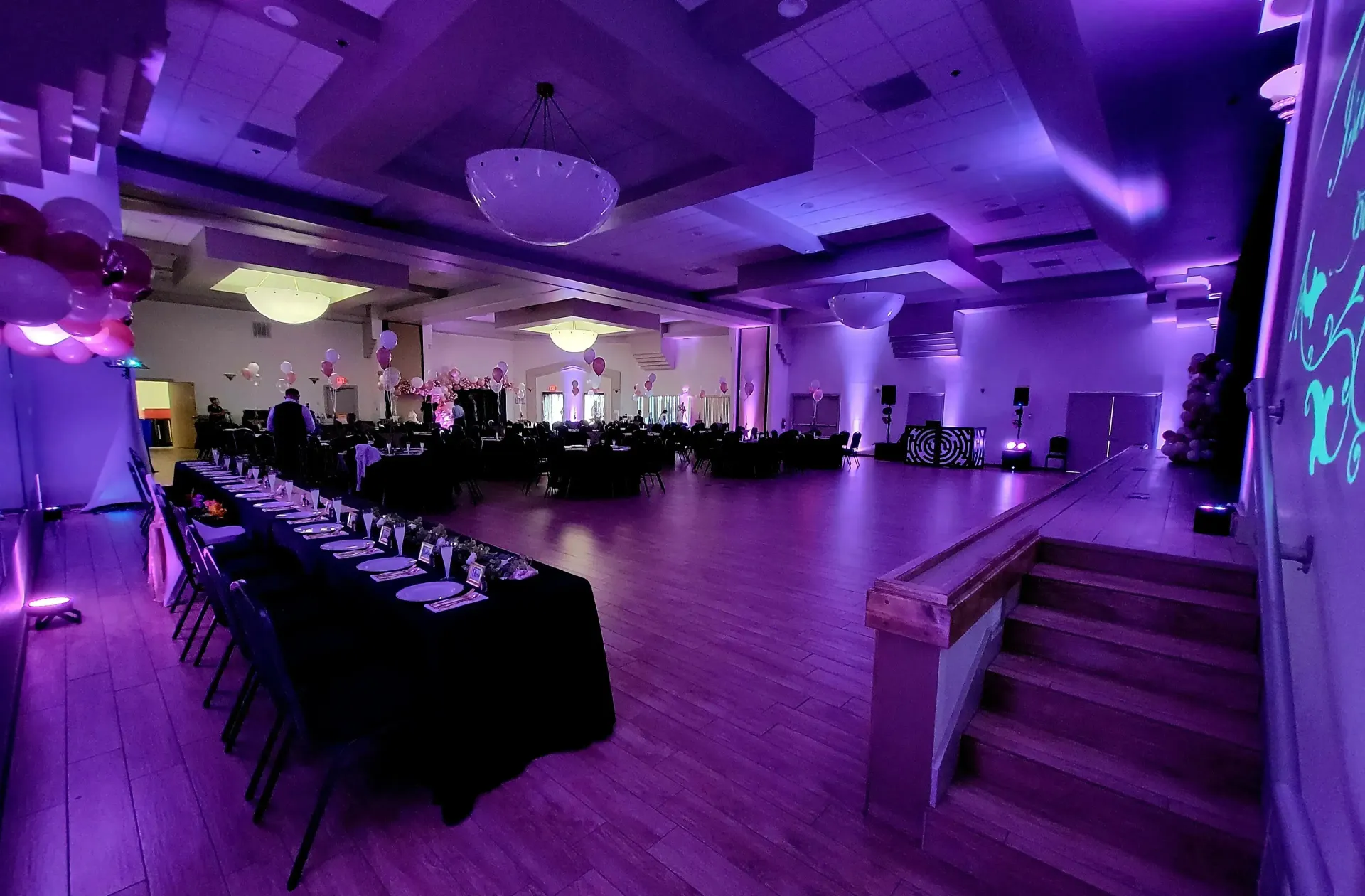 Purple-lit banquet hall with long tables, stage, and stairs leading up to a raised platform