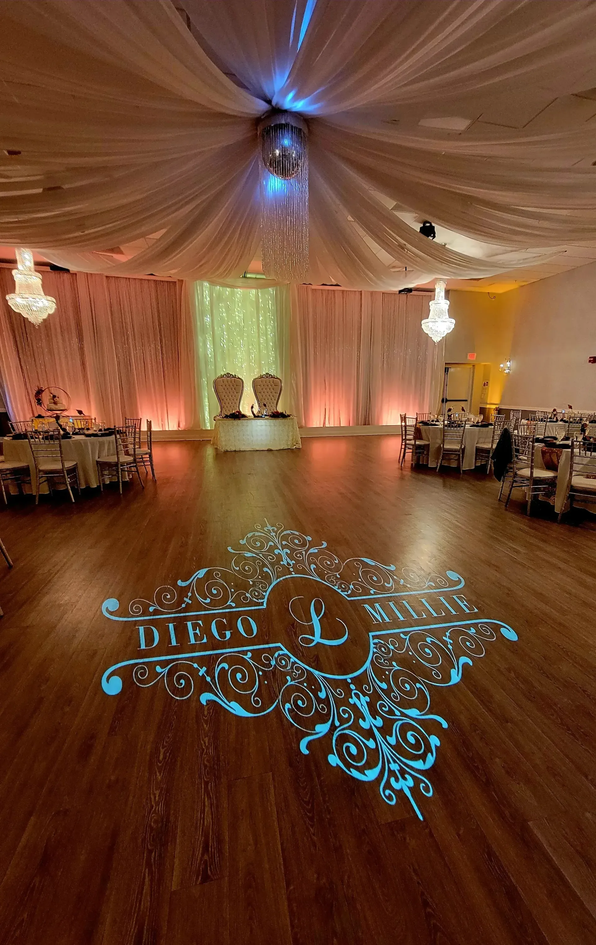 Elegant wedding reception hall with draped white ceiling, warm amber lighting, and a monogram projected on the floor