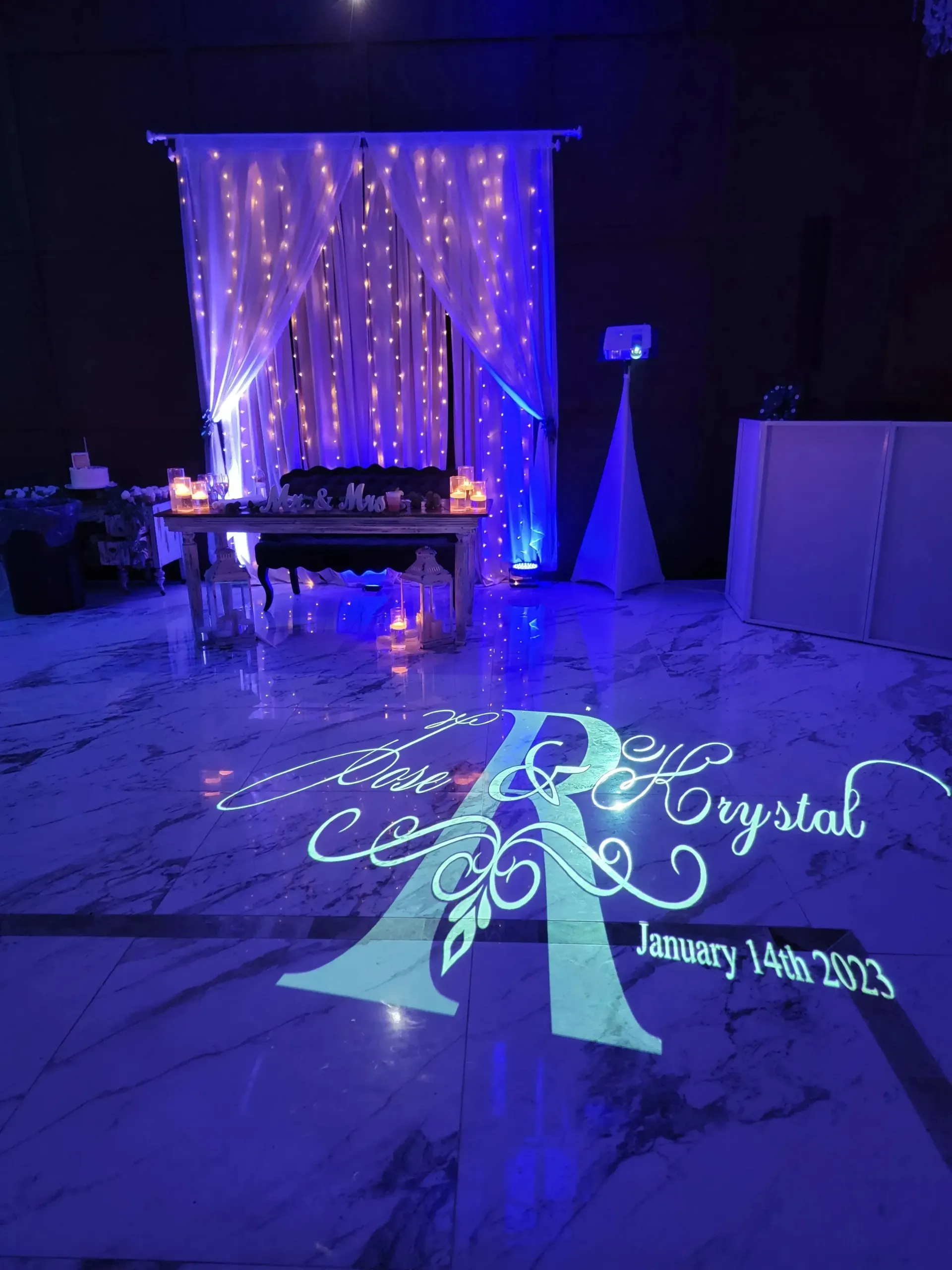 Blue-lit event room with a decorated table and projected monogram on the floor, with “Happy 140th, Crystal” text.