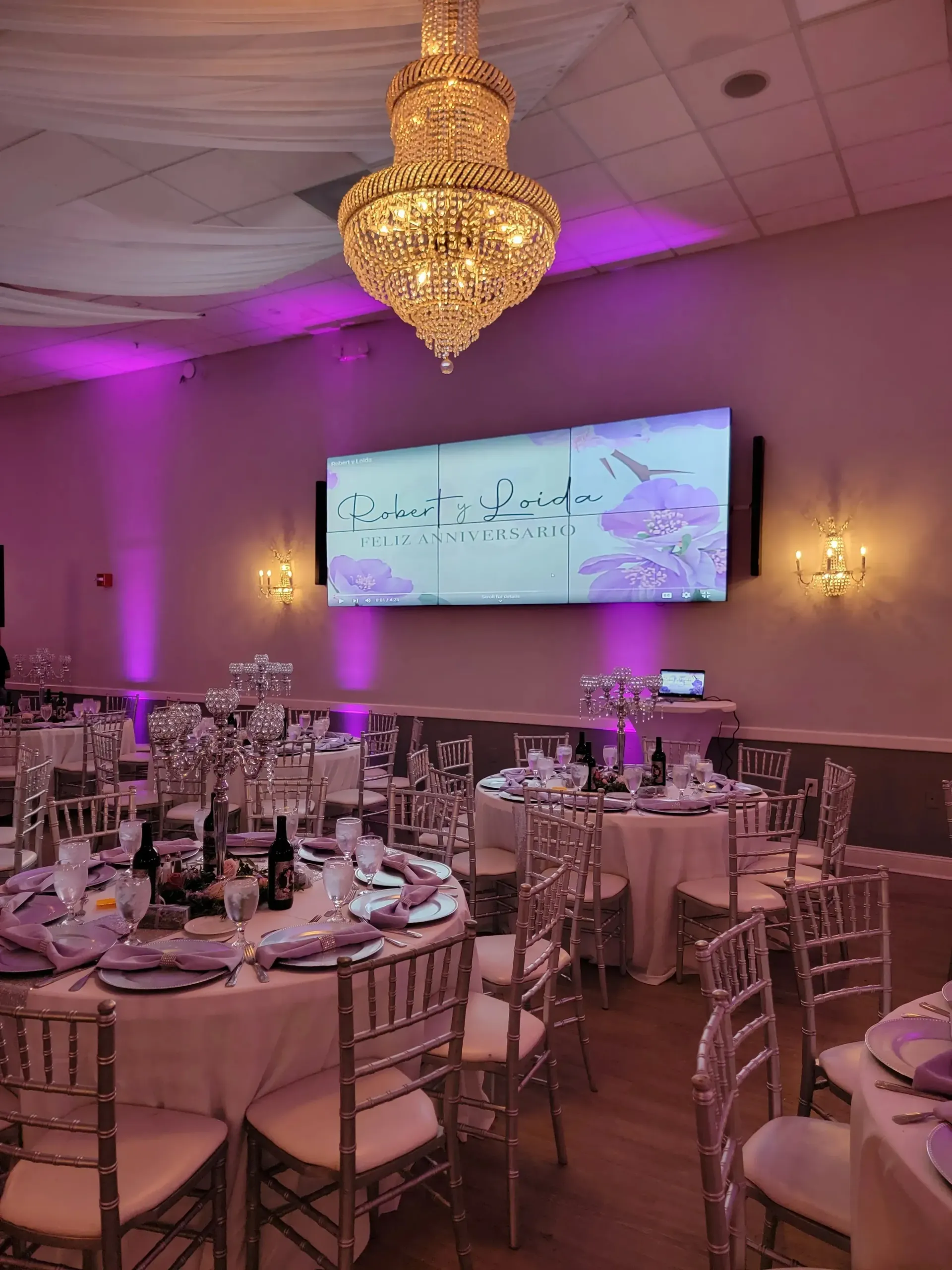 Elegant banquet hall with round tables, chandeliers, pink lighting, and a projection screen on the wall
