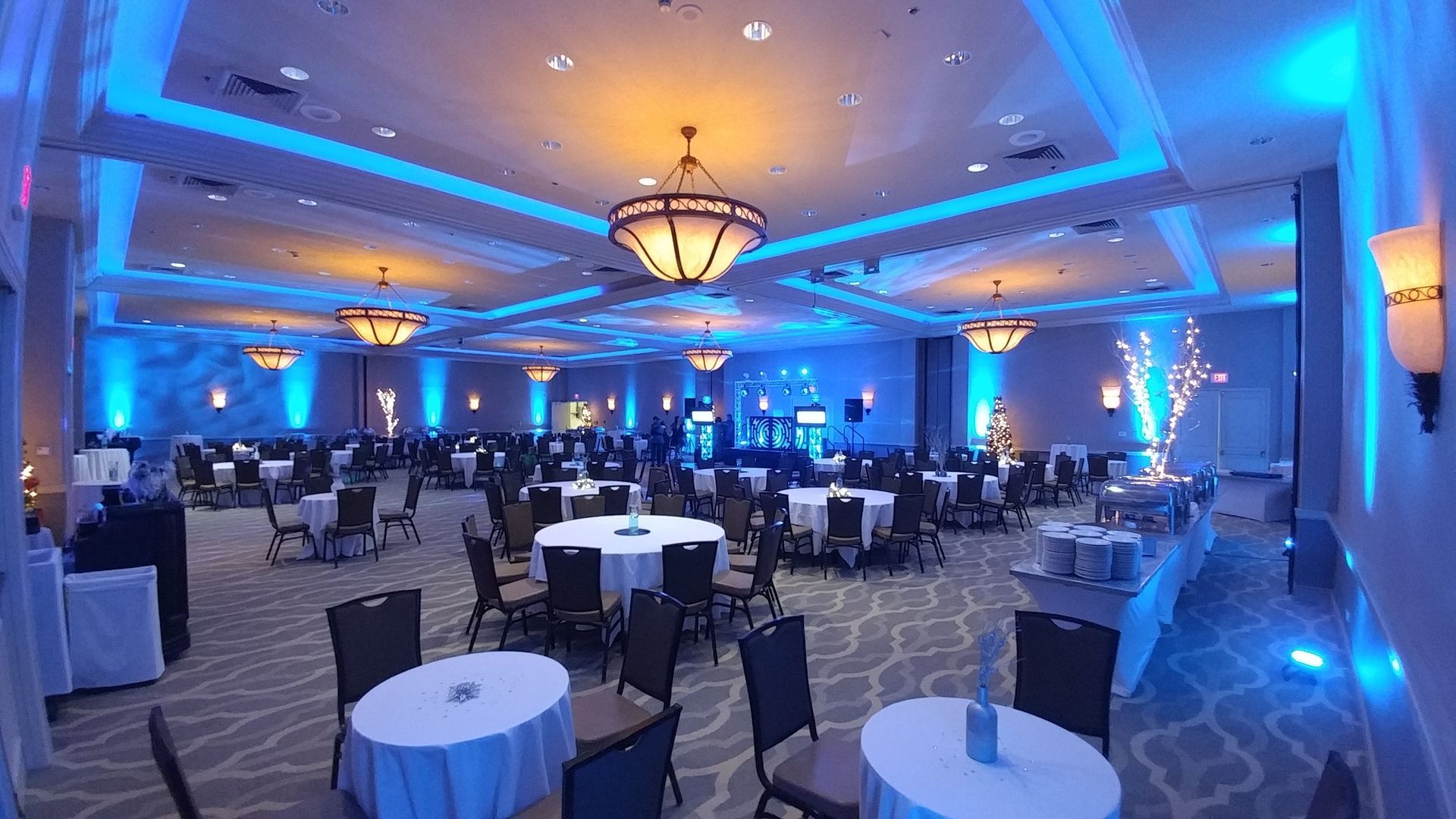 Blue-lit banquet hall with round tables, black chairs, and a large chandelier in the center.