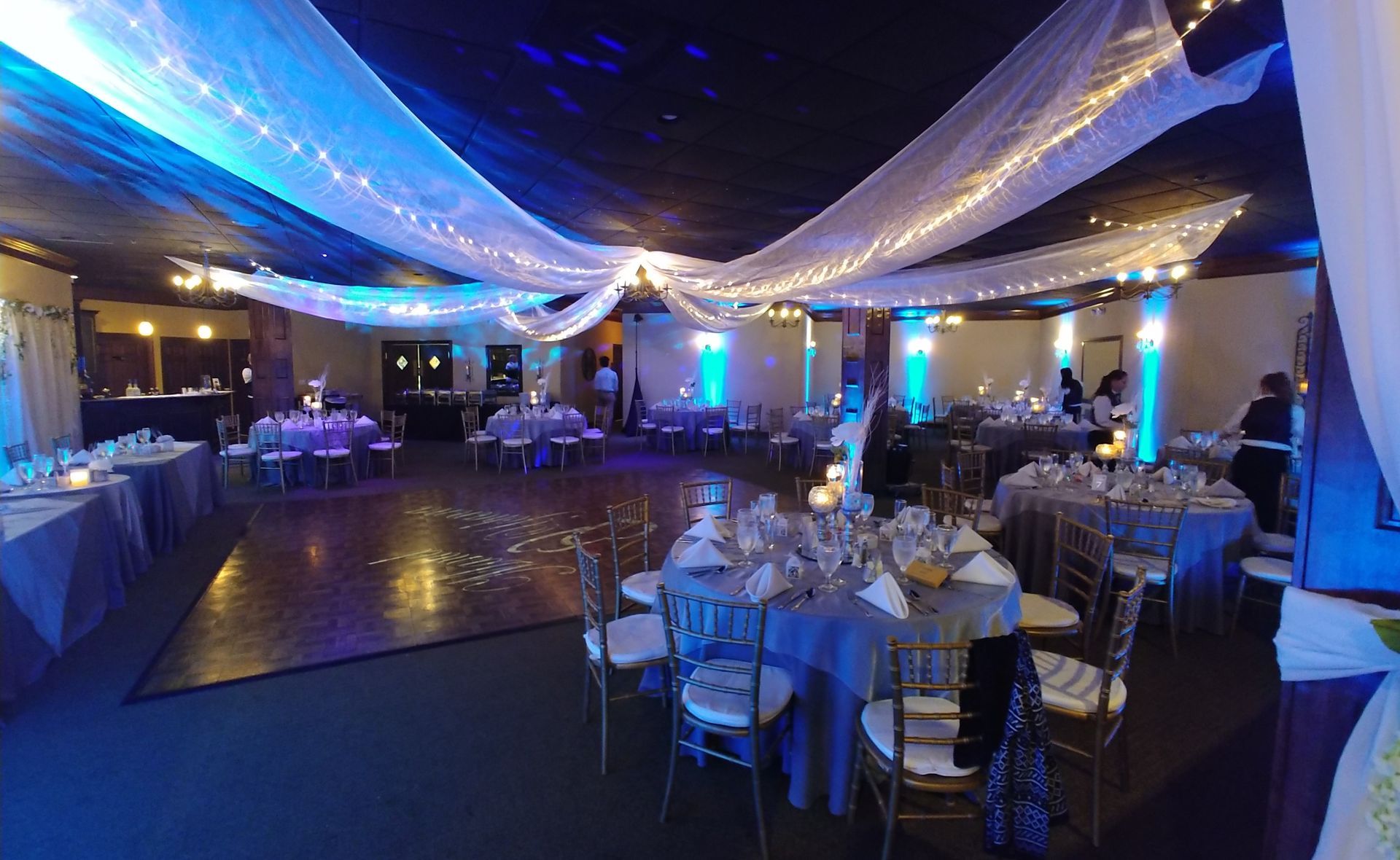 Elegant banquet hall with blue lighting, draped ceiling fabric, and set round tables for an event