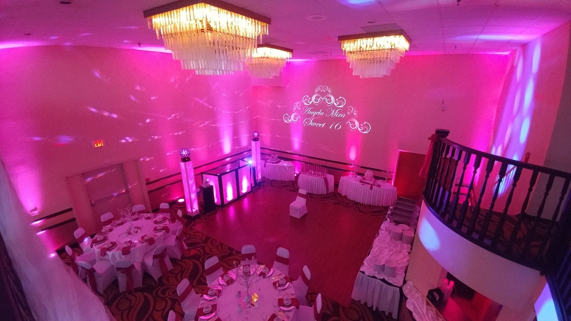 Pink-lit banquet hall with round tables, chairs, chandeliers, and a staircase on the right