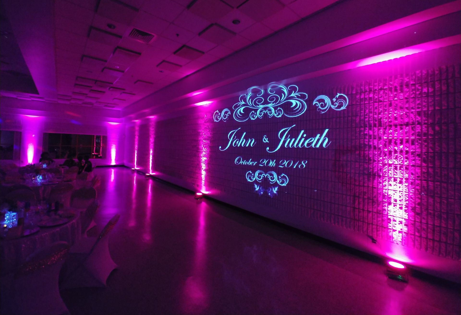 Purple-lit wedding reception hall with floral wall projection reading “John & Juliet”
