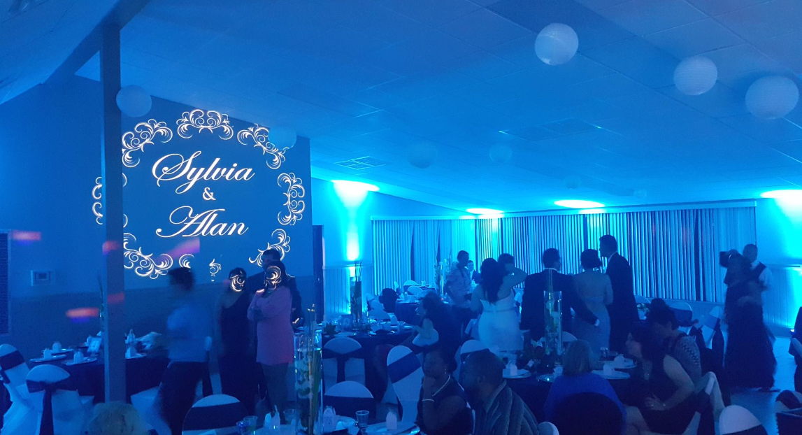 Blue-lit wedding reception with guests dancing and a lit sign reading “Sophia & Allan”