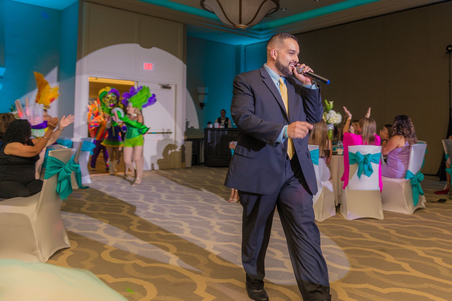 Man speaking at a banquet while colorful dancers perform in the background