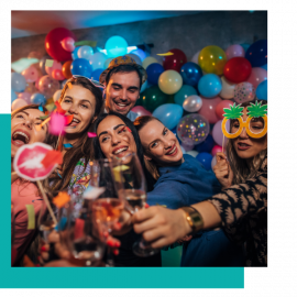 Group of friends smiling with drinks at a colorful balloon-filled party, posing for a selfie.