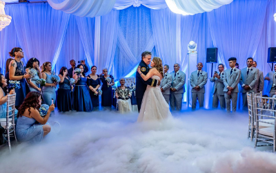 Bride and groom slow dancing under blue lights on a cloud-covered wedding floor, surrounded by guests