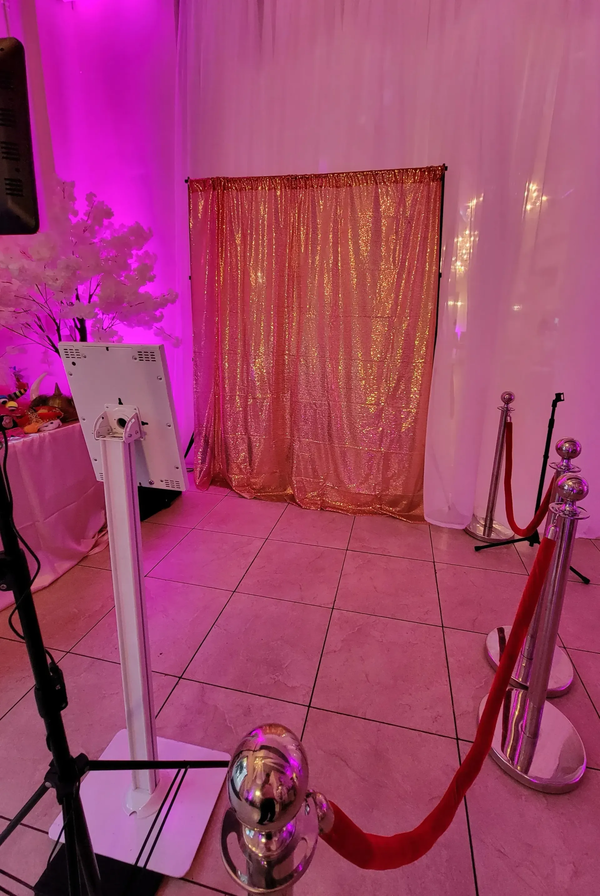 Pink-lit event backdrop with gold sequin curtain, rope stanchions, and a floral display on the left