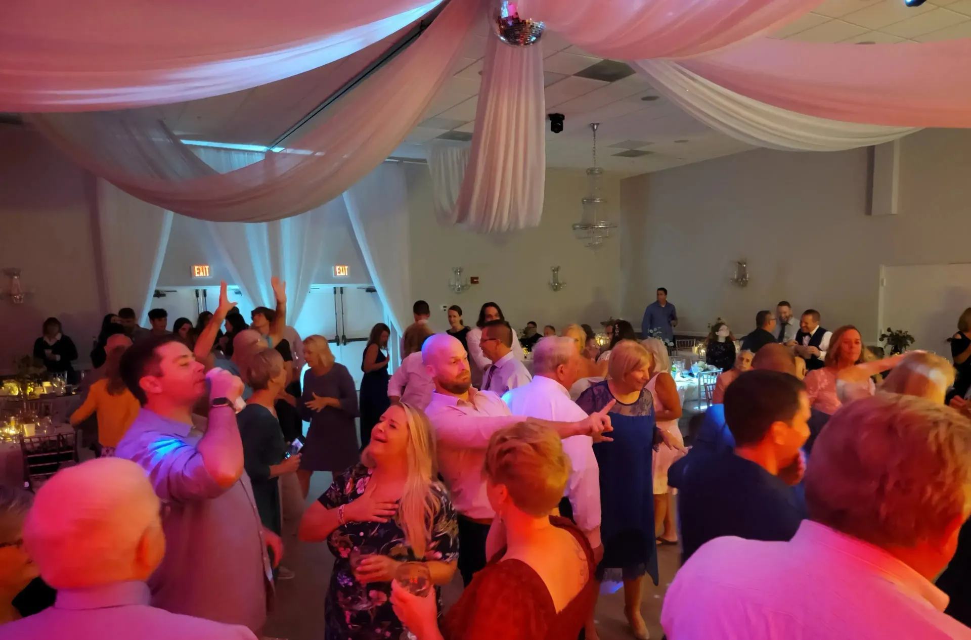 Crowded banquet hall with guests dancing under pink and blue lighting and draped ceiling decor