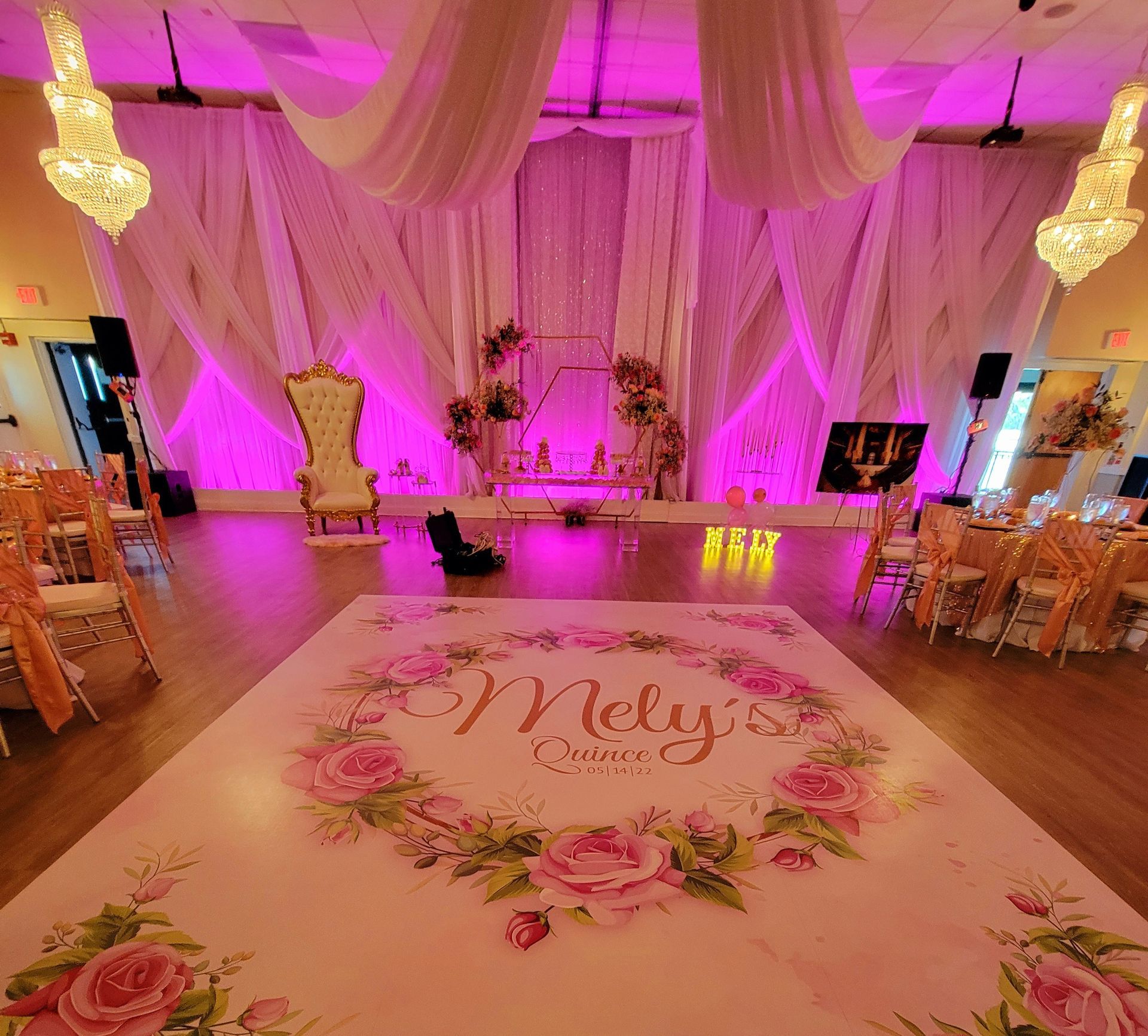 Pink-lit wedding reception hall with draped ceiling, floral decor, and dance floor monogram