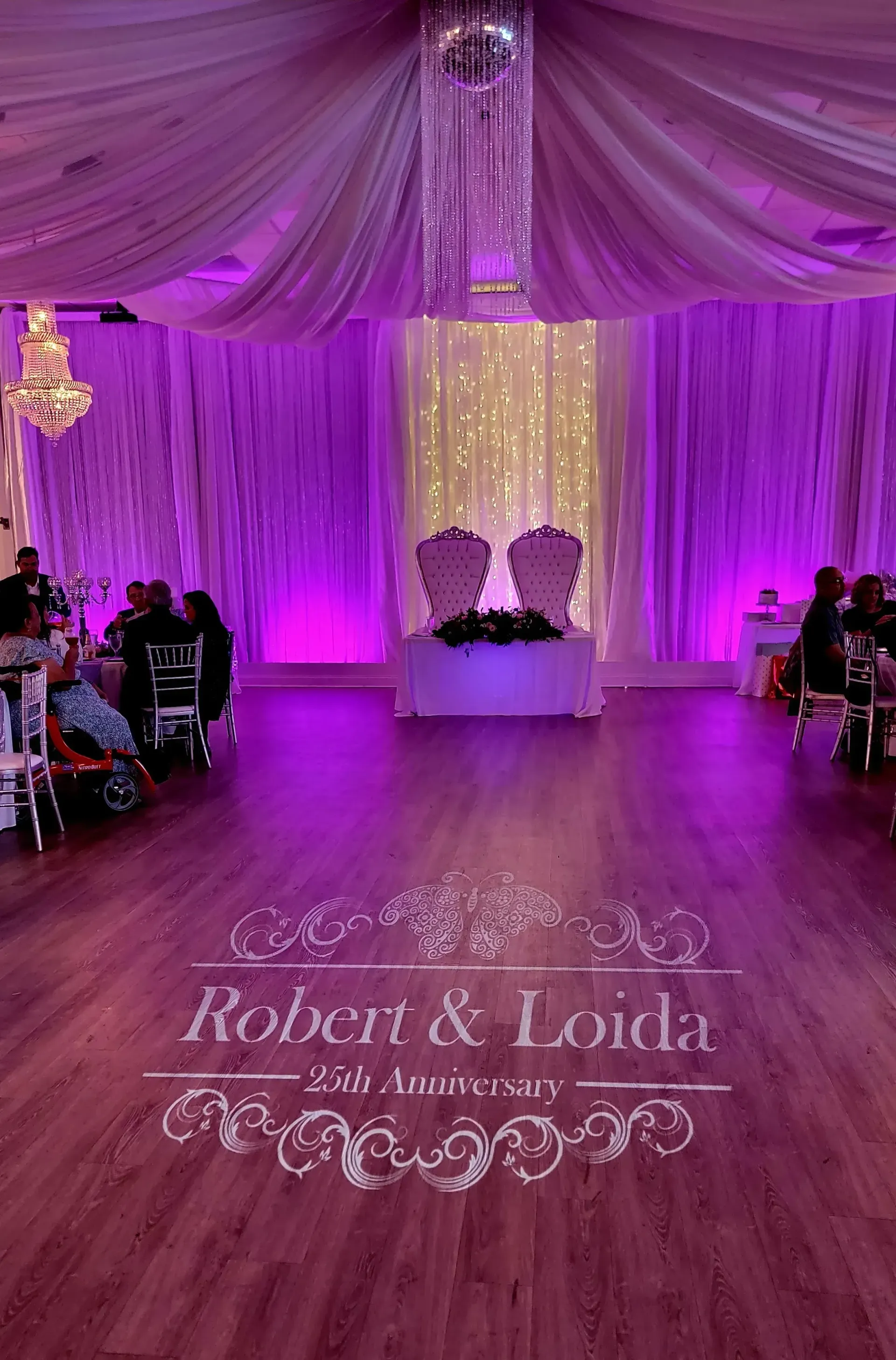 Purple-lit wedding venue with draped ceiling, dance floor, and white loveseat by a floral backdrop