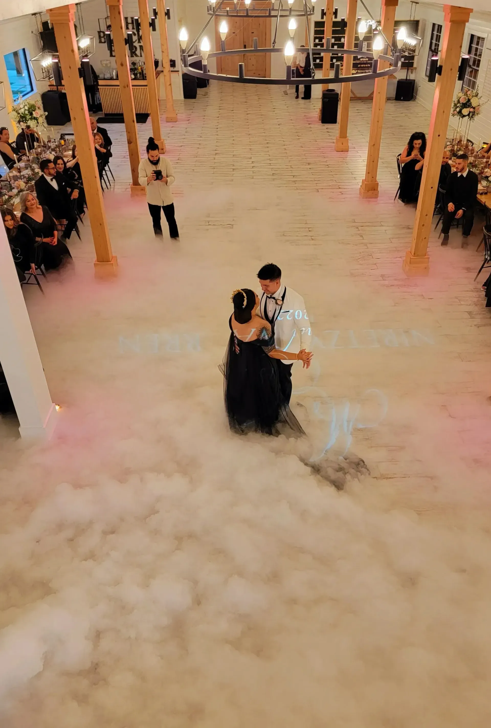 Couple dancing in a white-floored hall with fog, guests seated along both sides, warm lights overhead