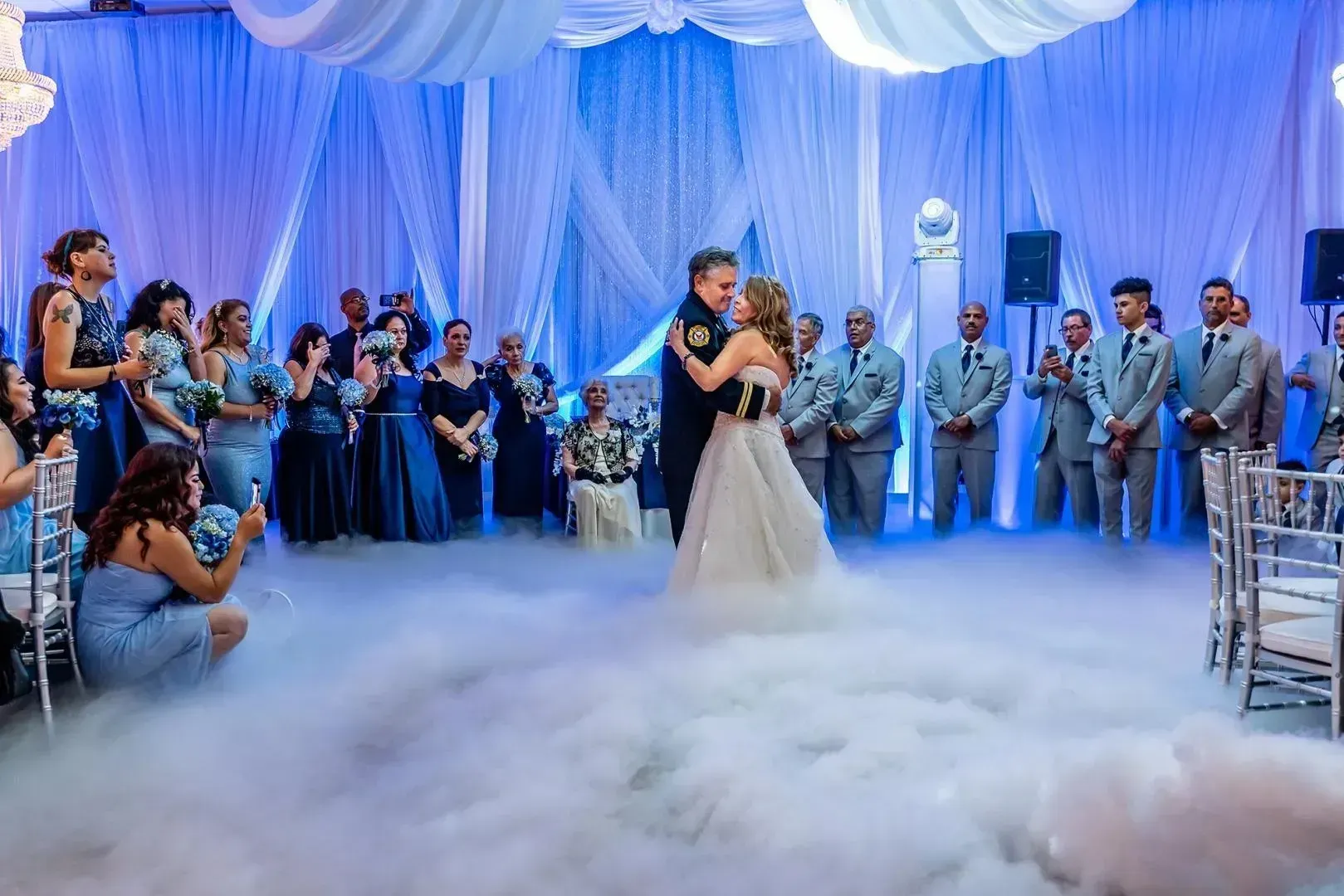 Bride and groom slow dance in a blue-lit wedding hall, surrounded by guests and low fog.