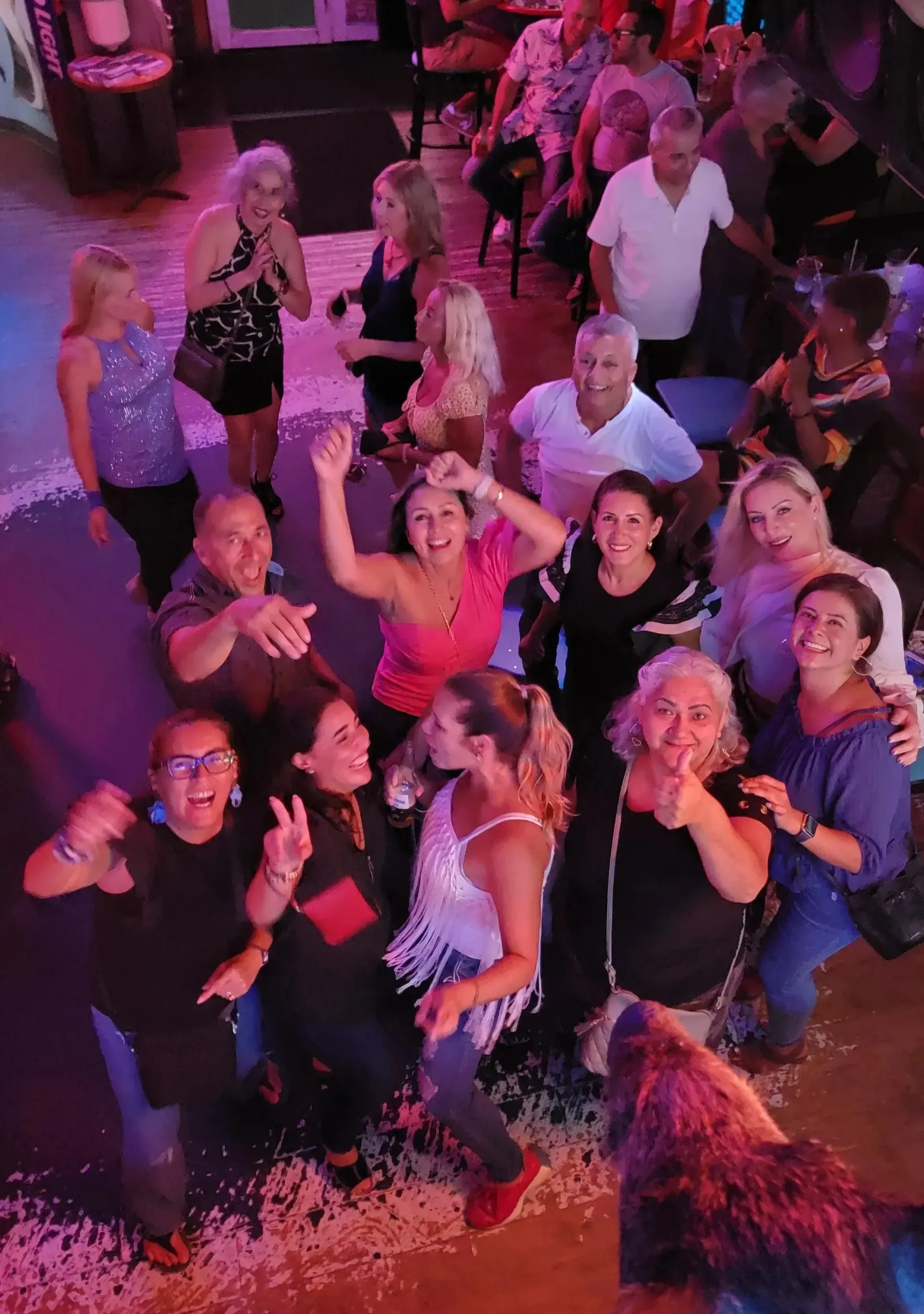 Group of people dancing and smiling under pink and purple party lights in a crowded club