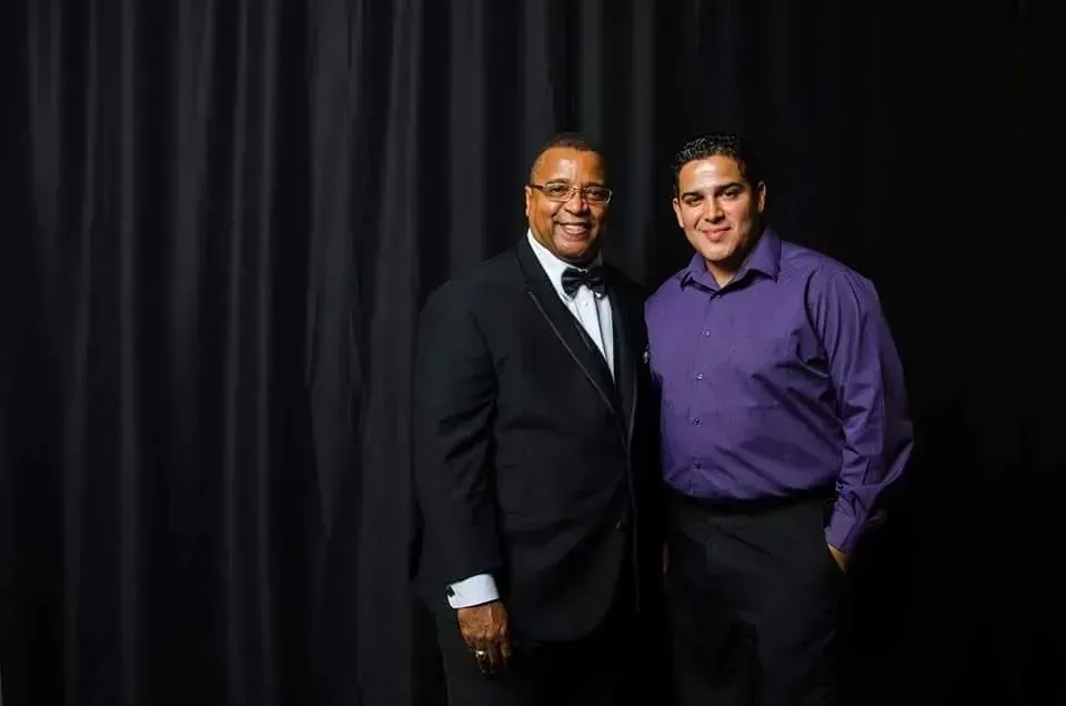 Two men standing together in formal attire in front of a dark curtain backdrop