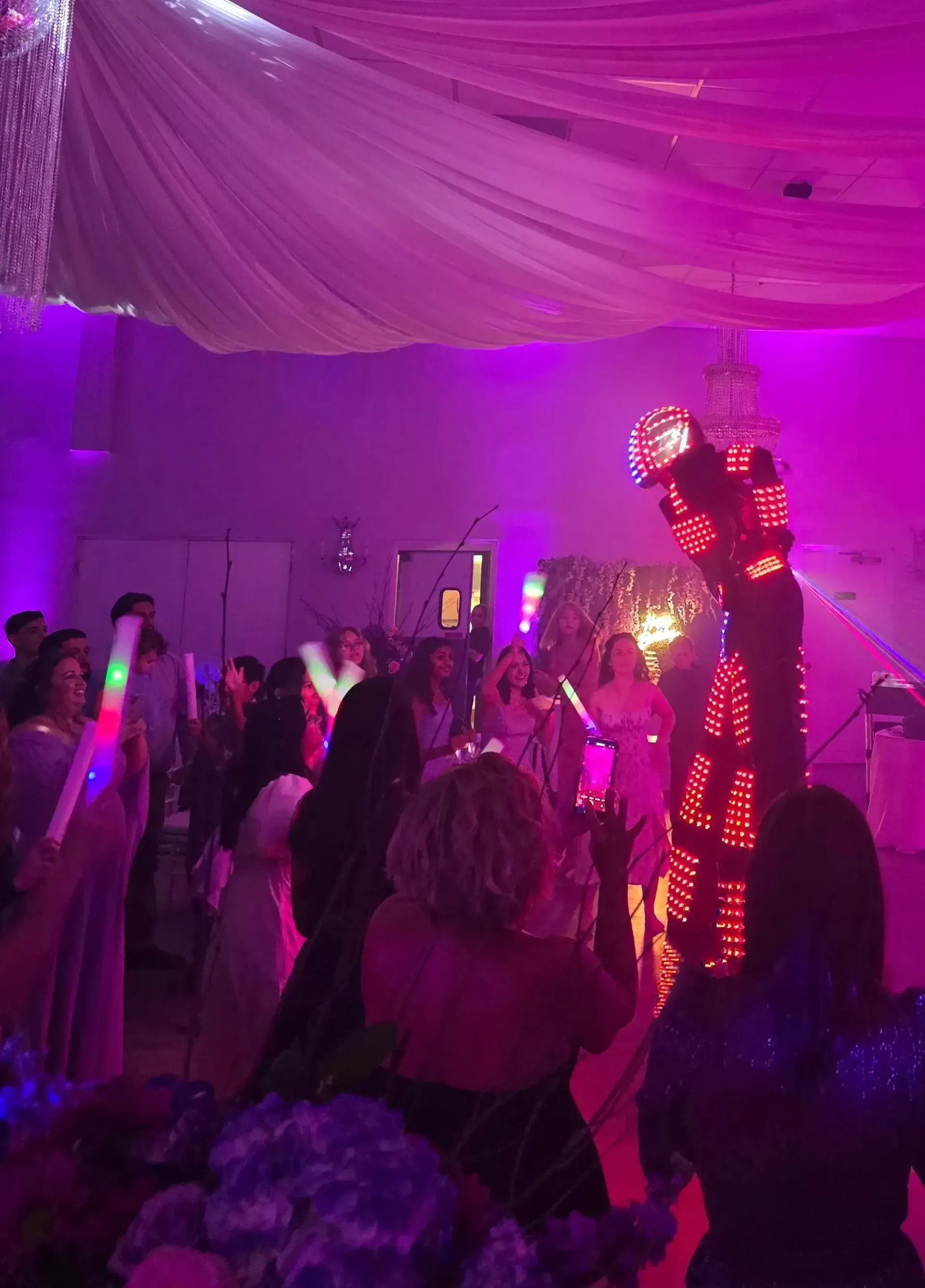 Crowded indoor party with purple lighting, draped ceiling fabric, and people dancing among glowing decorations