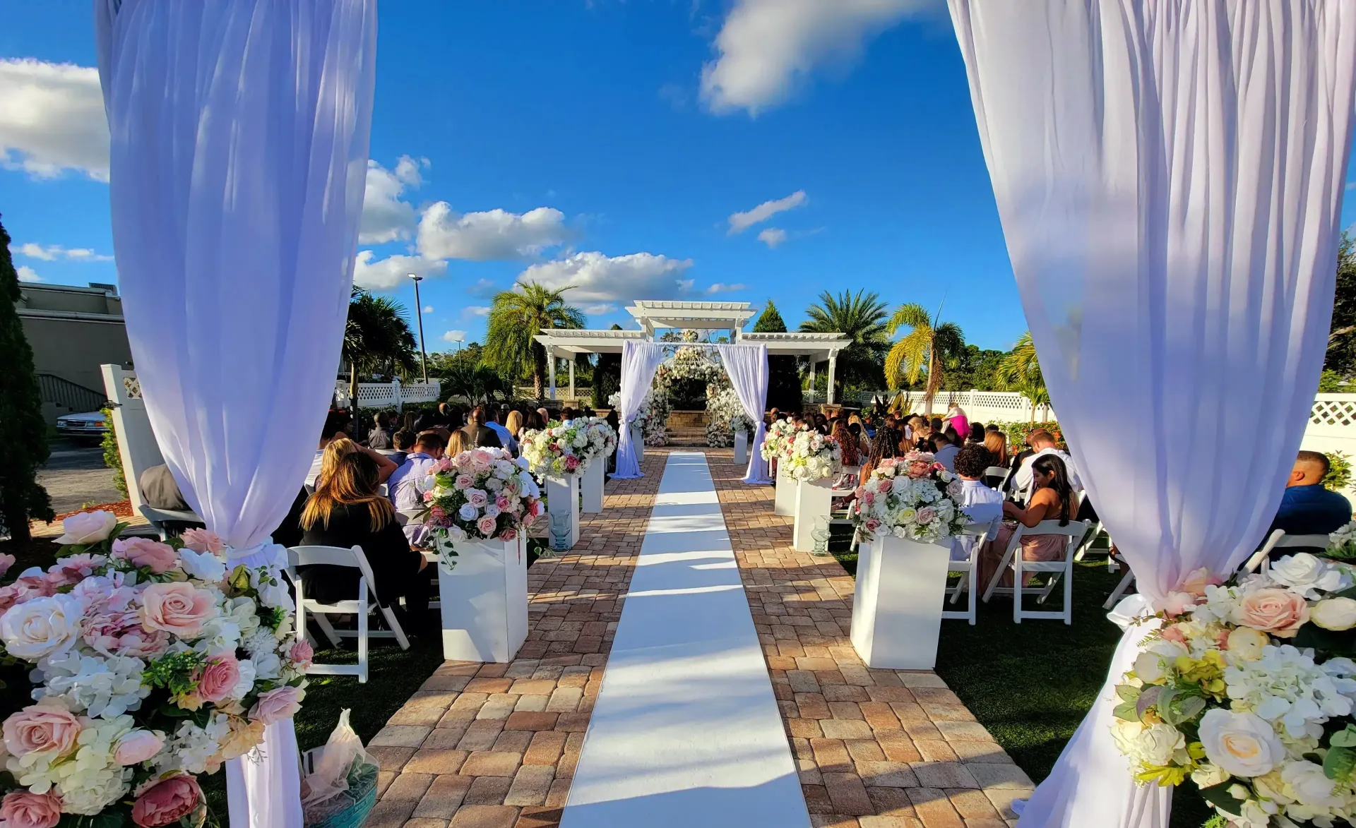 Outdoor wedding aisle with white drapes, floral arrangements, and guests seated under a blue sky