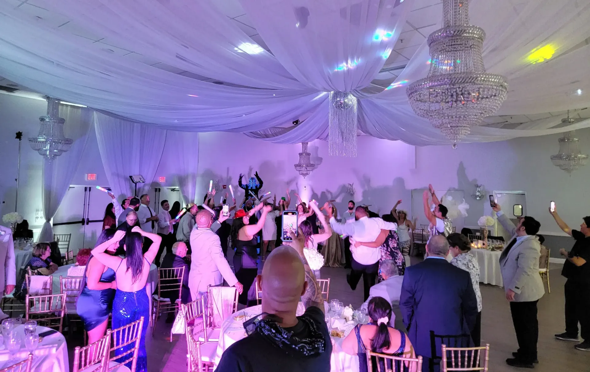 Guests dancing in a white-lit ballroom with chandeliers and draped ceiling during a formal celebration