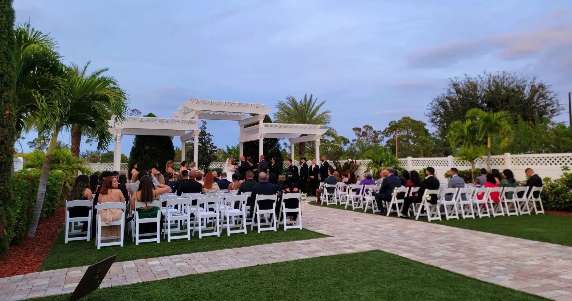 Outdoor wedding ceremony with seated guests, white chairs, and a floral arch on a lawn at sunset
