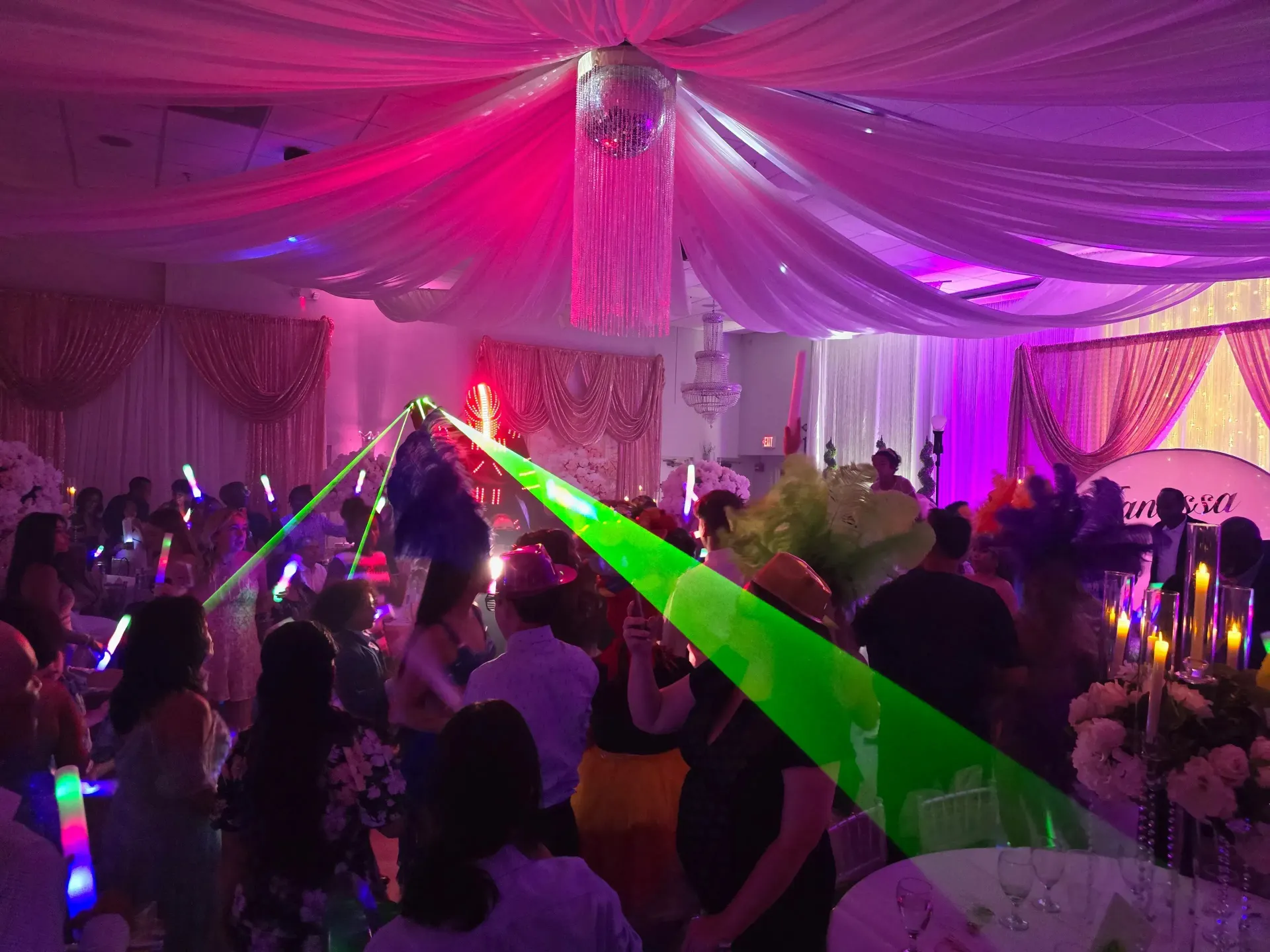 Crowded party tent with purple drapes and green laser lights, people dancing under chandeliers.