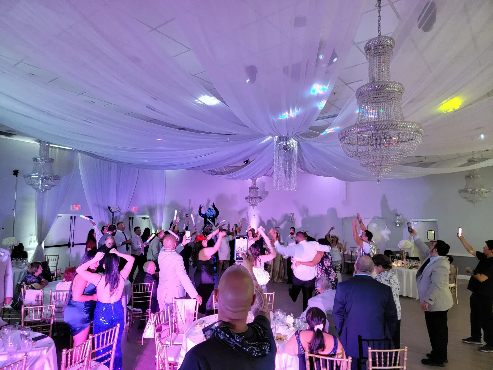 Guests dancing at a banquet hall under white draped ceilings and chandeliers, lit with purple and blue lights