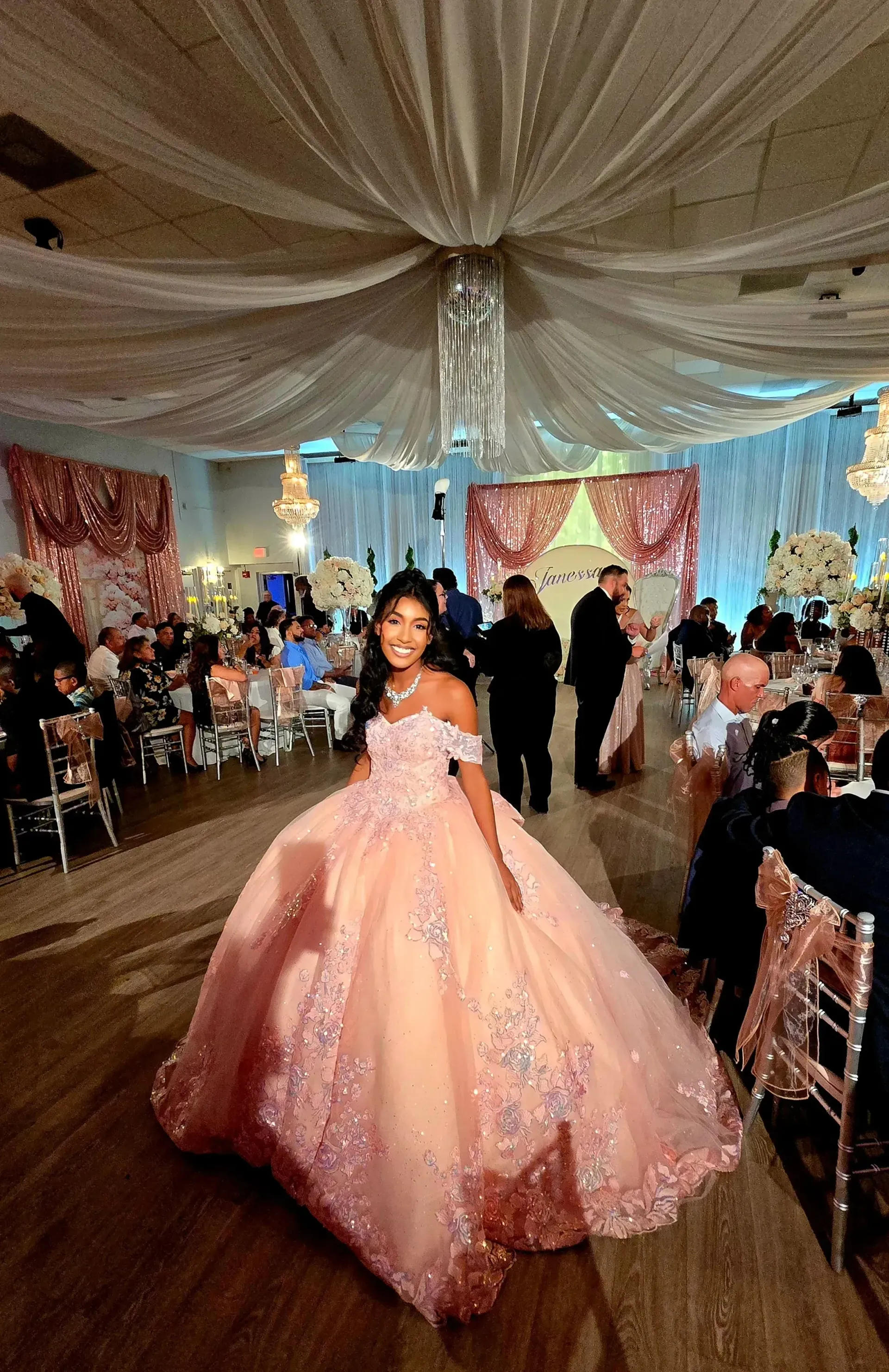 Young woman in a pink ball gown at a decorated indoor event hall with draped ceiling and seated guests
