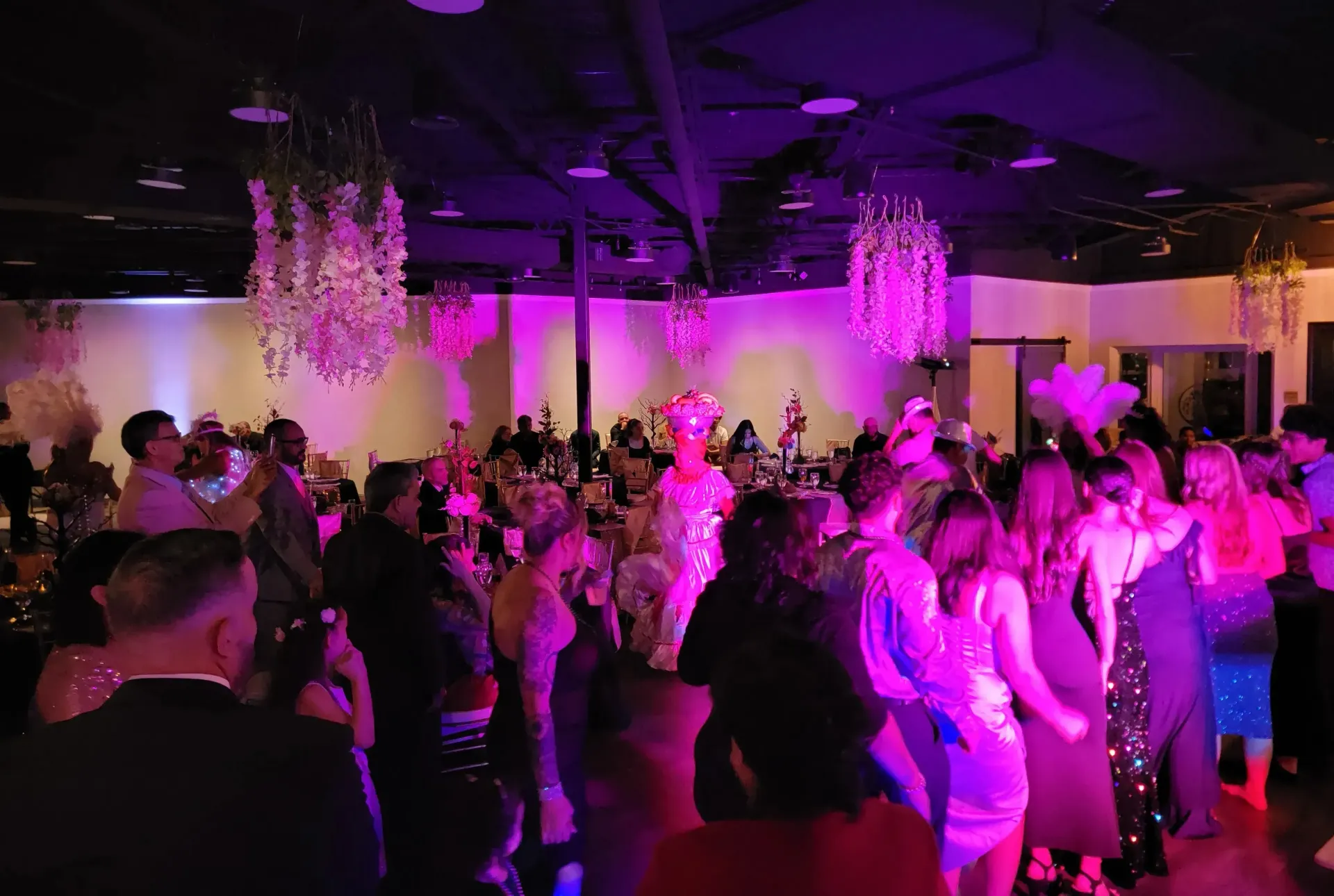Crowded ballroom dance floor lit in pink and purple with hanging floral decor and guests dancing