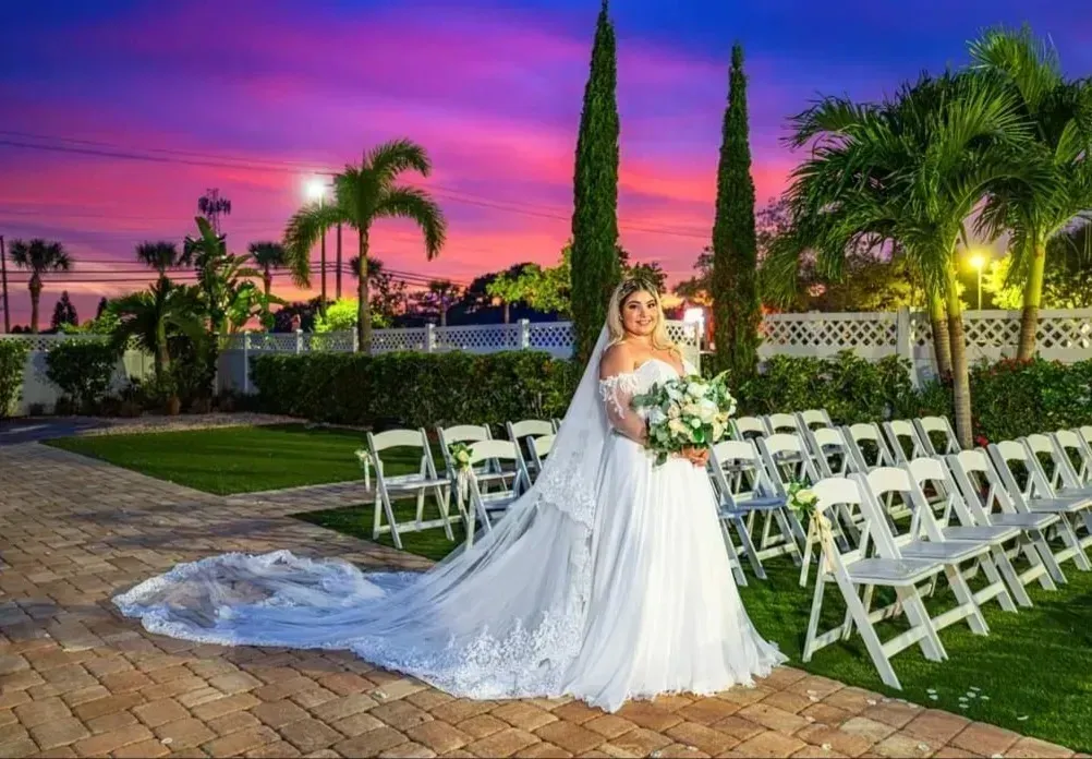 Bride in white gown holding bouquet at outdoor wedding venue at sunset, with rows of white chairs and palm trees