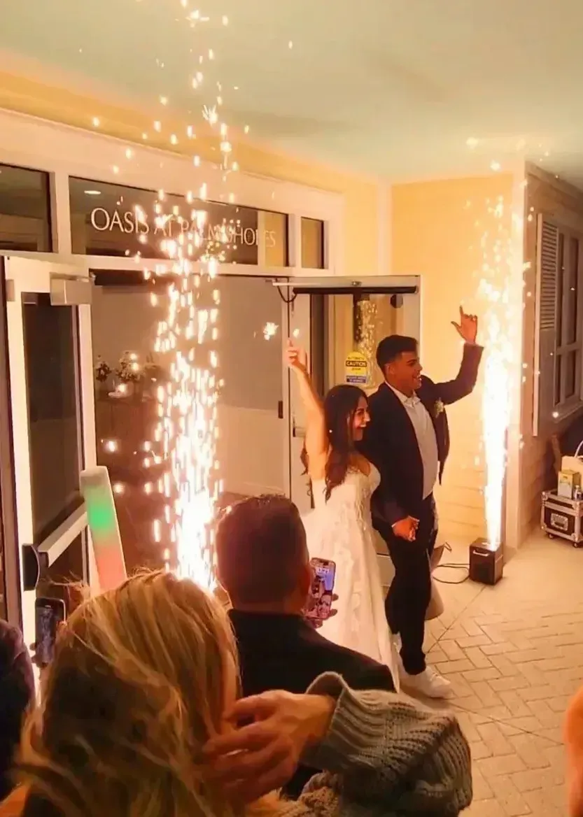 Wedding couple entering through indoor sparkler fountain, guests watching in a warmly lit room.