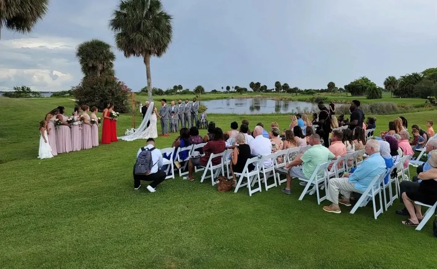 Outdoor wedding ceremony on a grassy lawn with guests seated and bridal party standing near an arch.