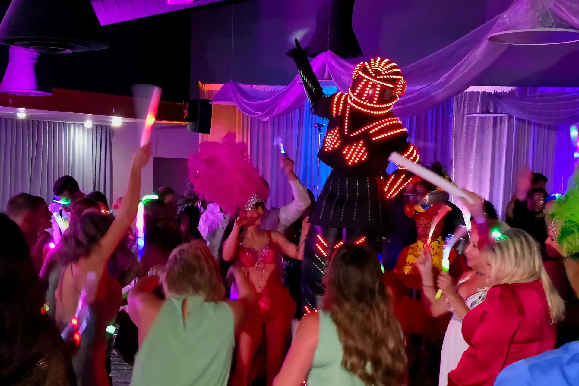 Crowd dancing at a neon-lit party while a performer in a glowing red mask stands on stage