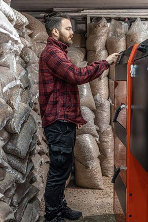 Man in red plaid Man in red plaid shirt operating a pellet boiler, surrounded by bags of wood pellets in a storage room.