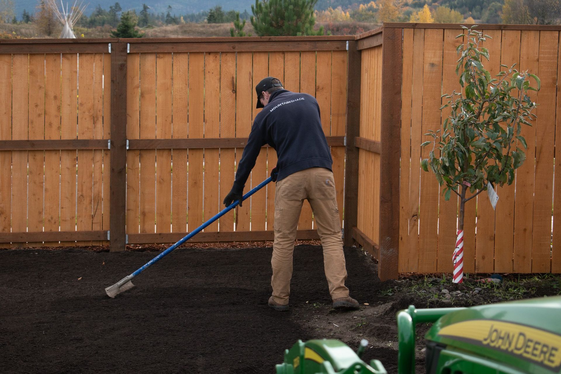 Person raking dark soil in a fenced yard beside a young tree and green John Deere equipment.