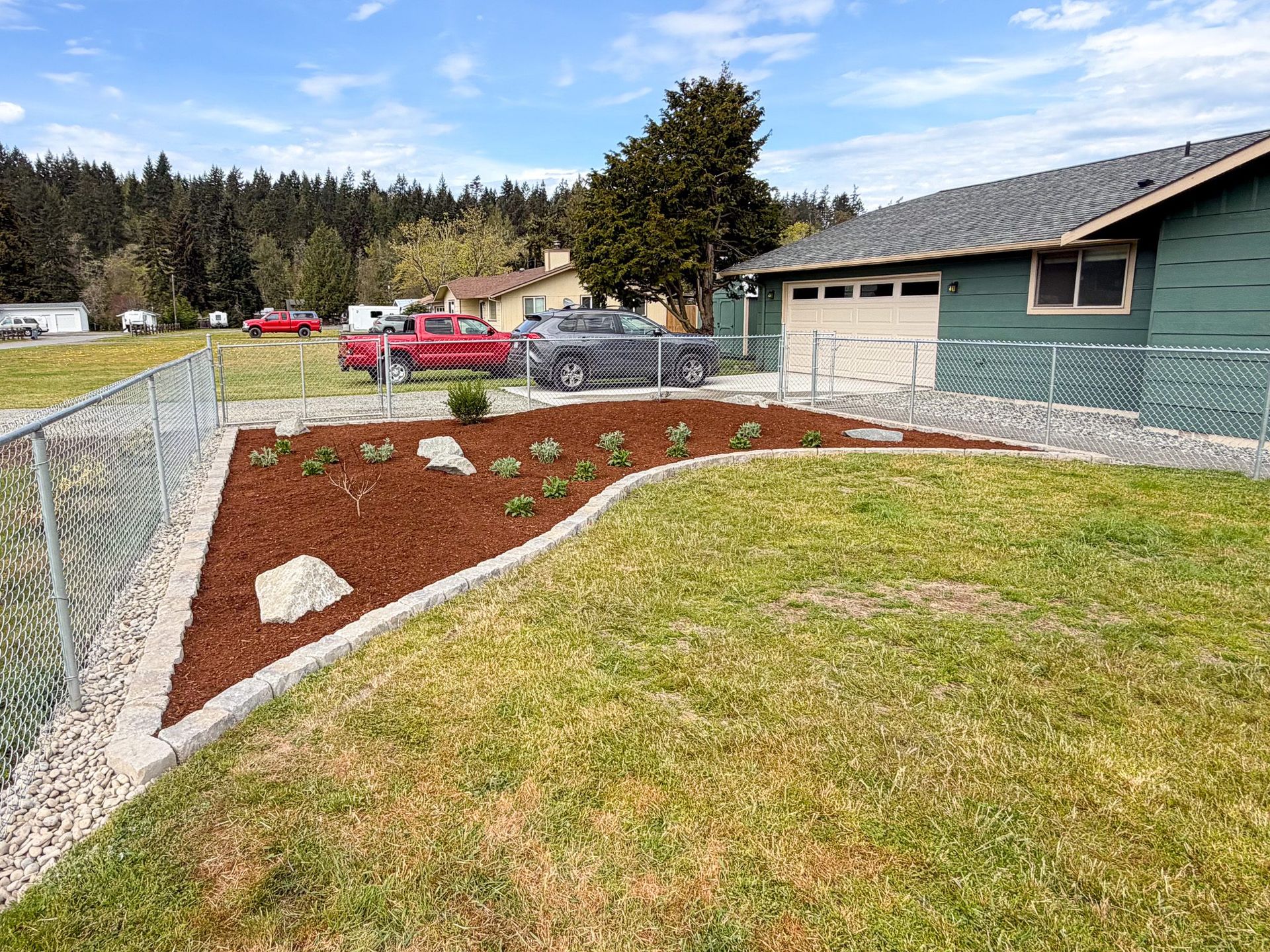 Landscaped yard with red mulch, curved grass edge, chain-link fence, and green building under a blue sky