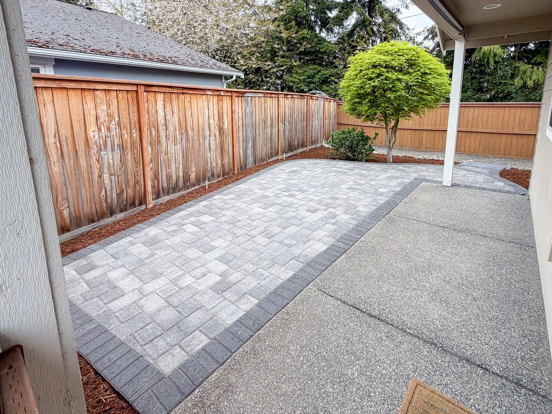 Backyard patio with gray pavers, a wooden fence, and a small tree near a covered area